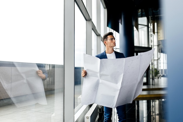 Architect in stijlvolle kleding houdt blad met tekening in zijn hand en praat via de telefoon op de achtergrond van een modern glazen gebouw met meerdere verdiepingen.