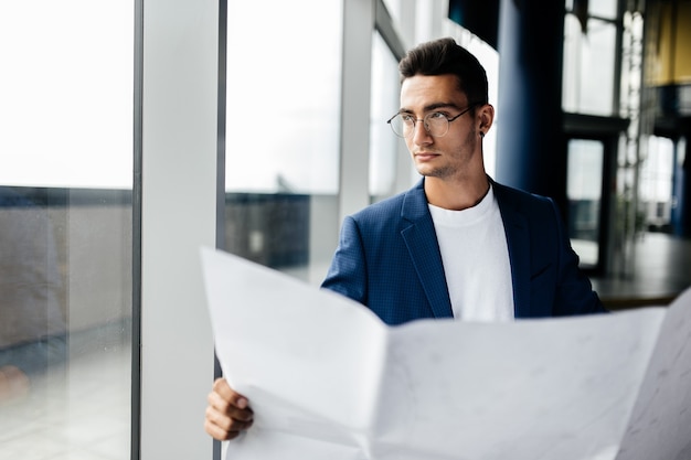 Architect in stijlvolle kleding houdt blad met tekening in zijn hand en praat via de telefoon op de achtergrond van een modern glazen gebouw met meerdere verdiepingen.