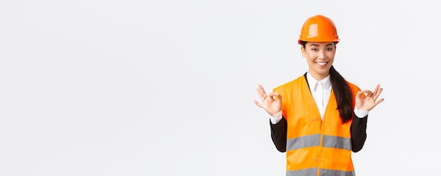 Architect made perfect plan Confident asian female engineer in safety helmet showing okay gesture have all under control pleased with excellent results standing white background