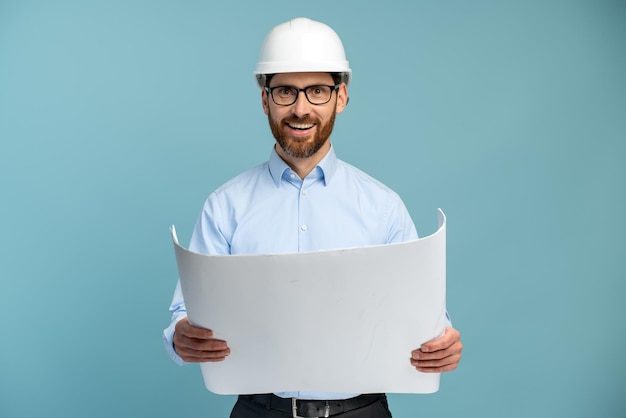 Architect man with cheerful face in builder safety helmet smiling while looking at the camera and holding blueprints over isolated background