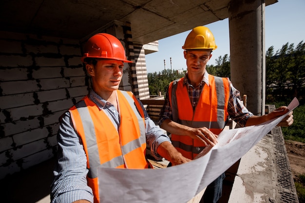셔츠, 주황색 작업 조끼 및 헬멧을 입은 건축가 및 구조 엔지니어는 건설 중인 건물 내부의 건설 현장에서 건설 문서를 탐색합니다.