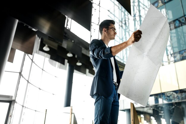 Architect in stylish clothes holds sheet with drawing in his hand and talks by phone on the background of a modern glass multistory building .