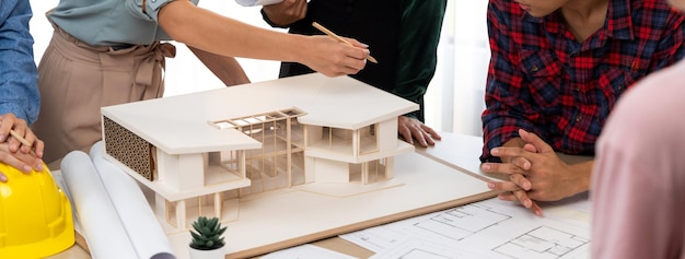 Architect team analysis and brainstorming about house construction at meeting table with house model blueprint and architectural equipment scatter around Creative design and teamwork Burgeoning