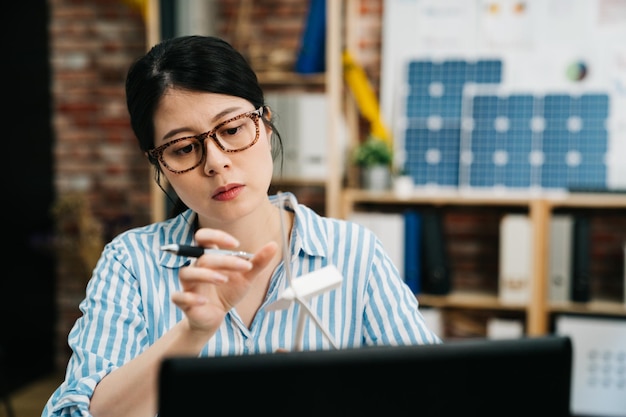 Architecture asian korean woman employee working at workspace in green office concept with red brick wall in background. interior designer eco friendly expert looking at windmill wind energy hold pen