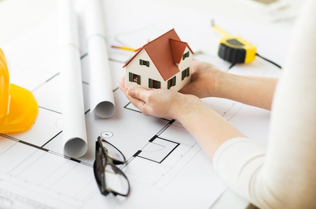 architecture, building, construction, real estate and people concept - close up of architect hands holding living house model above blueprint on table