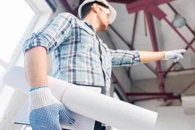 architecture and home renovation concept - man in helmet and gloves with blueprint showing direction