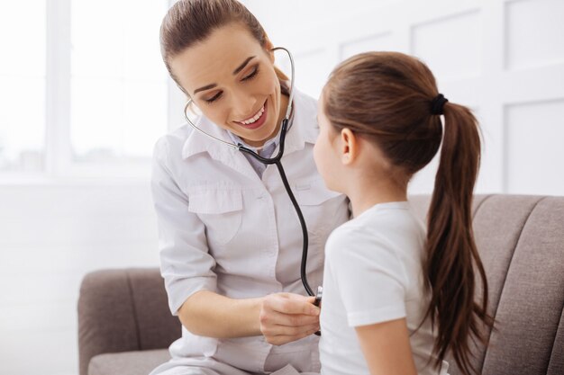 Are you ok. Pretty caring professional health worker consulting her little patient while listening to her breathing and looking delighted