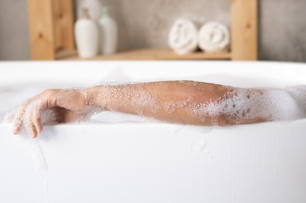 Рука молодого человека, расслабляющегося в белой фарфоровой ванне с пеной на полке с полотенцами и средствами по уходу за телом