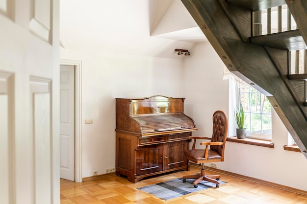 Кресло на ковре рядом с деревянным пианино в интерьере белой гостиной с растениями и лестницей Реальное фото