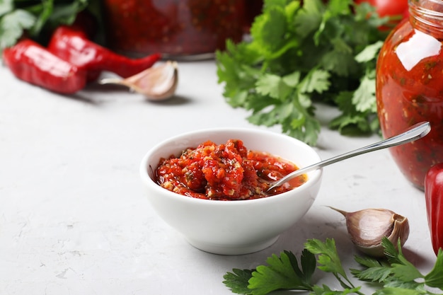 Armeense adjika van rode peper, tomaten, knoflook, koriander en peterselie in kom en potten, evenals verse ingrediënten op een tafel. Stilleven op grijze achtergrond.