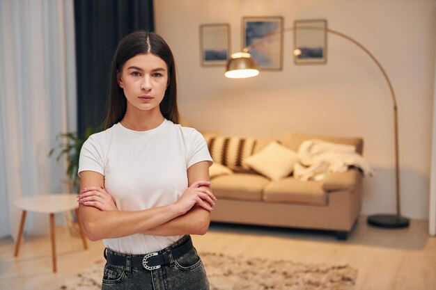 Arms crossed Woman in white shirt and black jeans is standing indoors at home