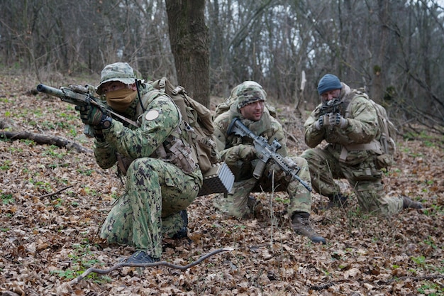 Army military soldiers team wearing camouflage uniforms and\
ammunition, armed assault rifles, machine gun replicas, hidings in\
woods, making ambush in forest, training in tactical teamwork