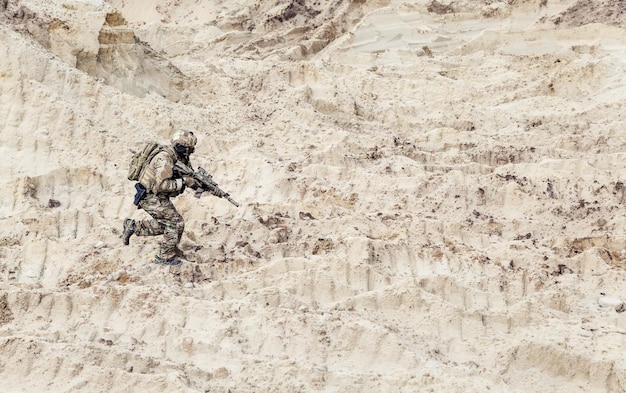 Army soldier or commando with assault rifle in ocp camouflage combat uniform, wearing helmet and with backpack on back running fast in sands. Special military operation in dessert, war on Middle East