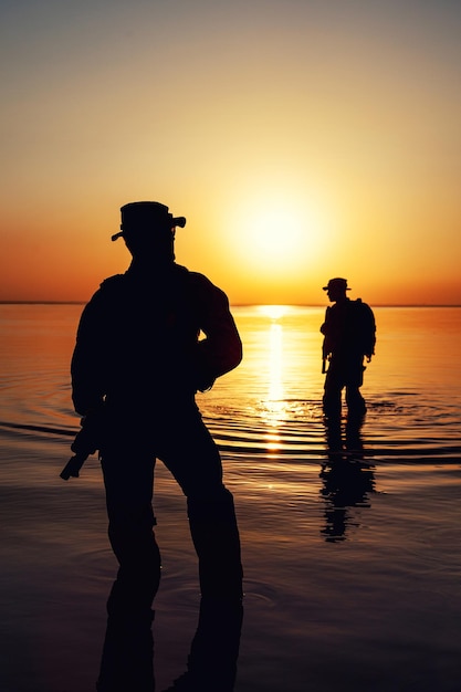 Army soldiers with rifles orange sunset silhouette in action during raid crossing river in the water