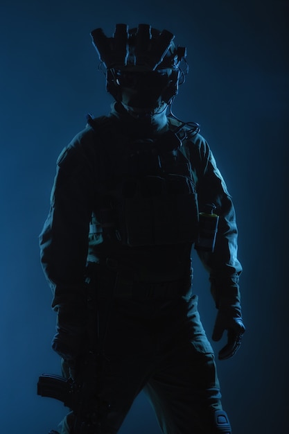 전술 탄약을 착용한 육군 특수부대 병사, 헬멧에 야간 투시경, 낮은 키에 낮은 서비스 라이플을 들고 서 있는, 백 뷰, 패브릭 배경에 빨간색 백라이트가 있는 스튜디오 촬영