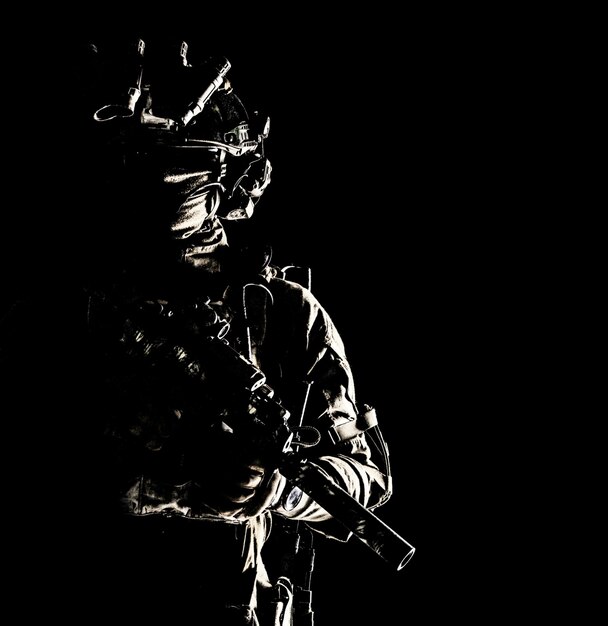 Army special operations forces soldier in mask and combat uniform helmet equipped nightvision device armed submachine gun with silencer looking aside low key studio portrait on black copyspace