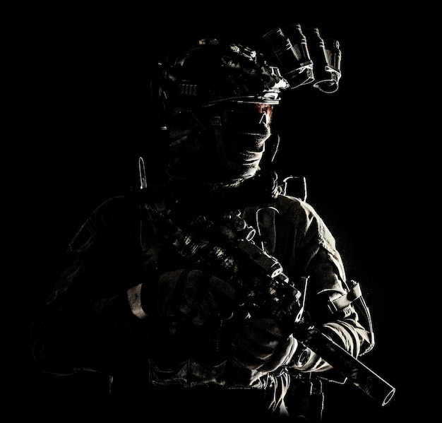 Army special operations forces soldier in mask and combat uniform helmet equipped nightvision device armed submachine gun with silencer looking aside low key studio portrait on black copyspace