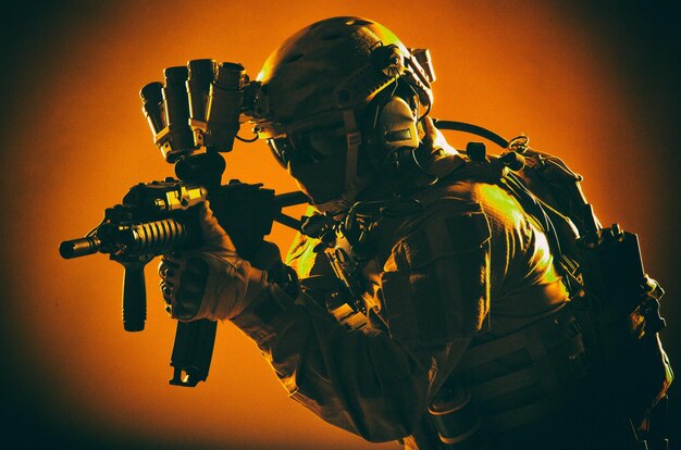 Army special operations soldier, commando fighter in full tactical ammunition, helmet with radio headset and night vision device, aiming short barrel assault rifle in darkness, low key studio shoot