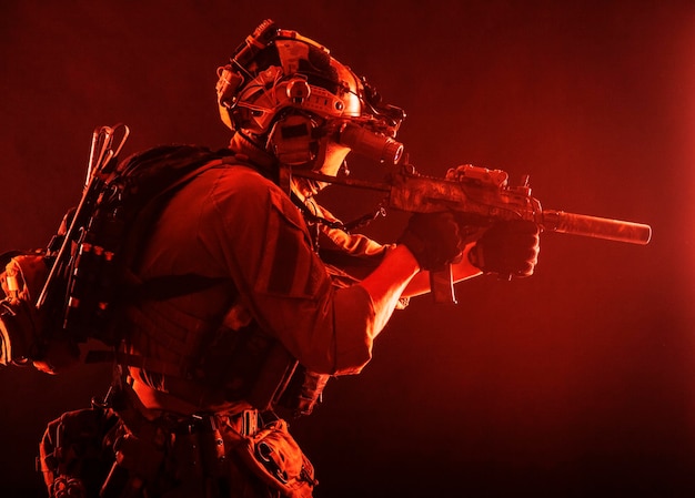 Photo army special operations soldier commando fighter in full tactical ammunition helmet with radio headset and night vision device aiming short barrel assault rifle in darkness low key studio shoot