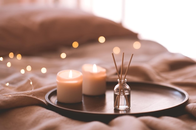 Aroma bamboo sticks in bottle with scented liquid with candles staying on wooden tray in bed closeup.