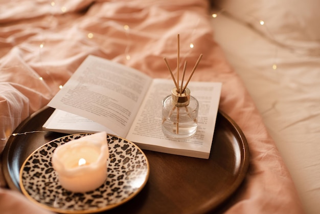 Aroma sticks with liquid scented perfume water in bottle with burning candle staying on open paper book on wooden tray in bed close up Aromatherapy Home cozy atmosphere