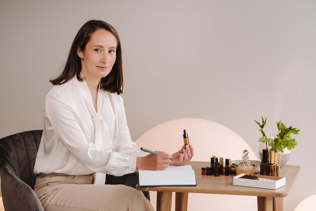 The aromatherapist girl is sitting in her office and holding a bottle of aromatic oil in her hands and writing something down there are essential oils on the table