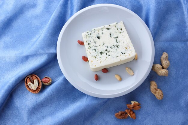 Aromatic cheese with blue mold and nuts on a white plate on a blue background Closeup of a slice of dorblu cheese