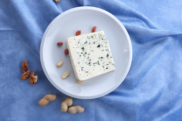 Aromatic cheese with blue mold and nuts on a white plate on a blue background Closeup of a slice of dorblu cheese