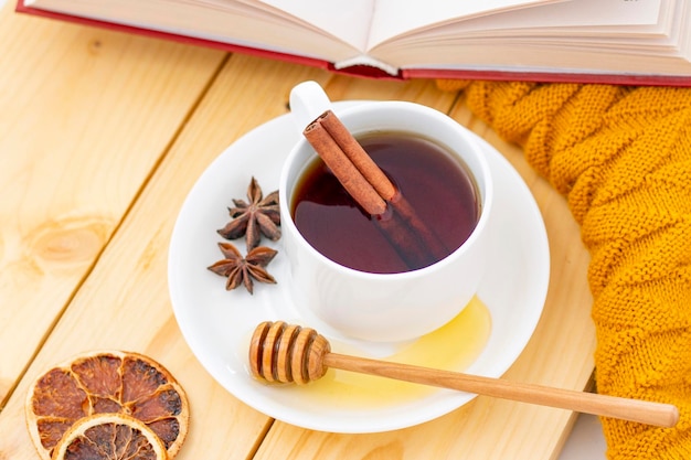 Aromatische hete kaneelthee bedekt met een warme sjaal op een houten herfstachtergrond, honingdipper met honing, comfortabel om een boek te lezen