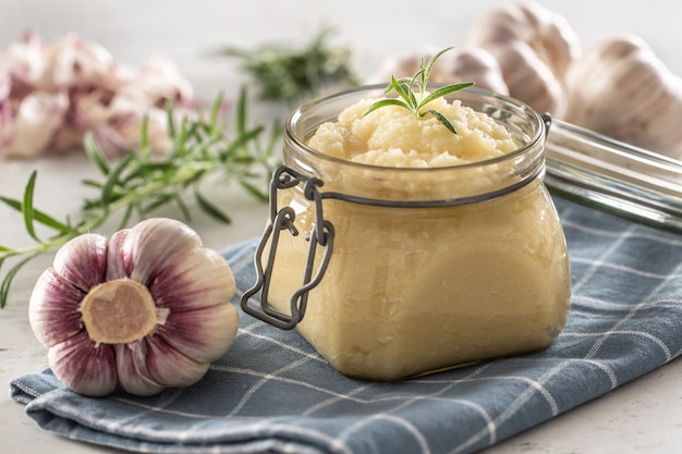 Foto aromatische knoflookpasta in een glazen pot op rustieke keukendoek met bollen en gepelde kruidnagel en rozemarijn.