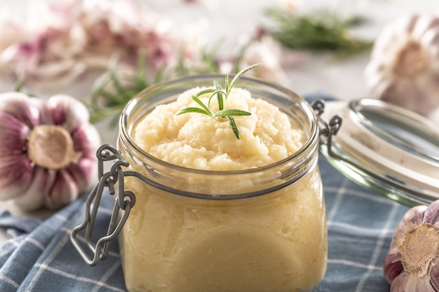 Aromatische knoflookpasta in een glazen pot op rustieke keukendoek met bollen en gepelde kruidnagel en rozemarijn.