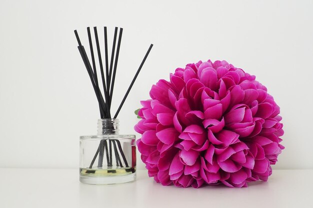 Arrangement with incense sticks essential oil in a glass vase and a bright pinklilac terry artificial flower in the interior of a white room White background