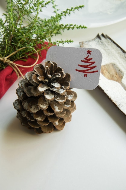 Arranging the table for the winter holidays concept. Table decoration in minimal style, red napkin with herbs, the outline of a dessert spoon, pine cone and holiday place card