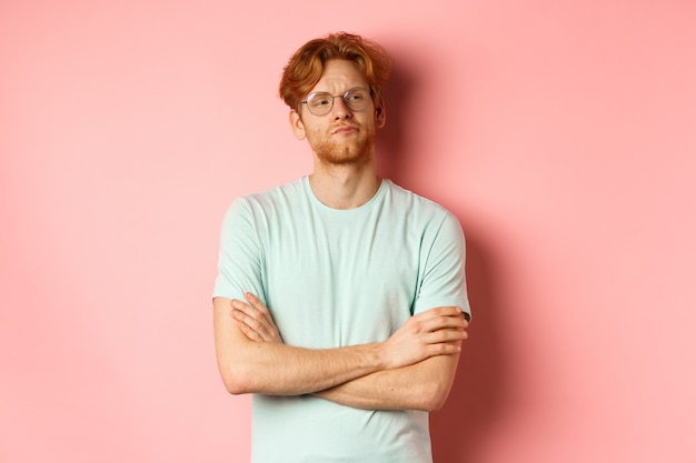 Arrogant redhead guy in glasses cross arms on chest, looking at something with skeptical face, standing over pink background.