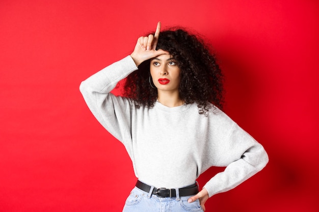 Arrogante vrouw met krullend haar, verliezersteken op voorhoofd tonen en opzij kijken, iemand bespotten, staande op rode achtergrond.