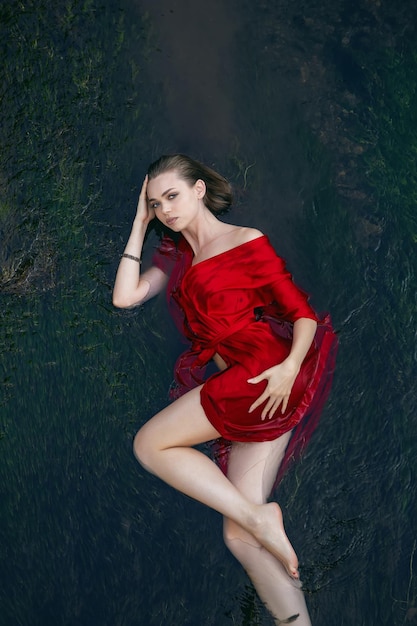 Art beautiful romantic portrait of a sexy young woman in a red dress lying in a river with green algae in summer in nature during the day