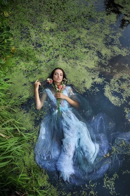 Искусство красивая романтическая женщина лежит в болоте в синем длинном платье с цветами. Брюнетка портрета в прозрачном платье в ряске грязи болота воды. Книжная обложка