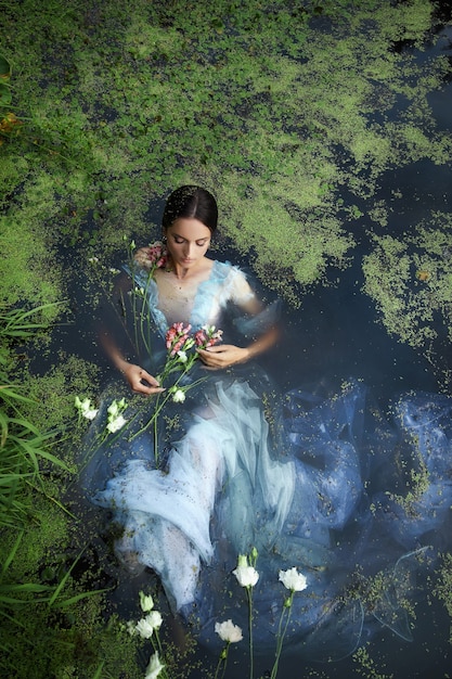 Arte bella donna romantica si trova nella palude in abito lungo blu con fiori. bruna del ritratto in vestito trasparente in lenticchia d'acqua di fango di palude d'acqua. copertina del libro
