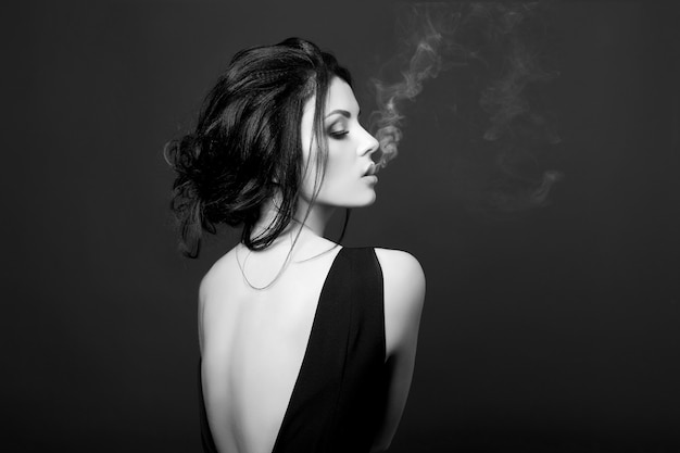 Art Brunette woman Smoking on dark background in black dress. Classic portrait of confident strong woman