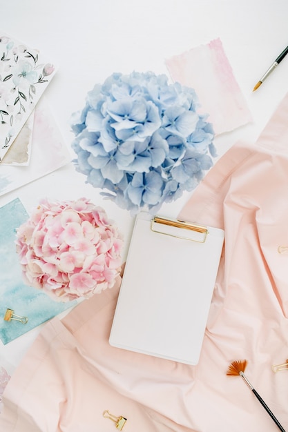 Art composition home office desk workspace with copy space clipboard, pastel hydrangea flowers bouquet, watercolor painting, peachy blanket