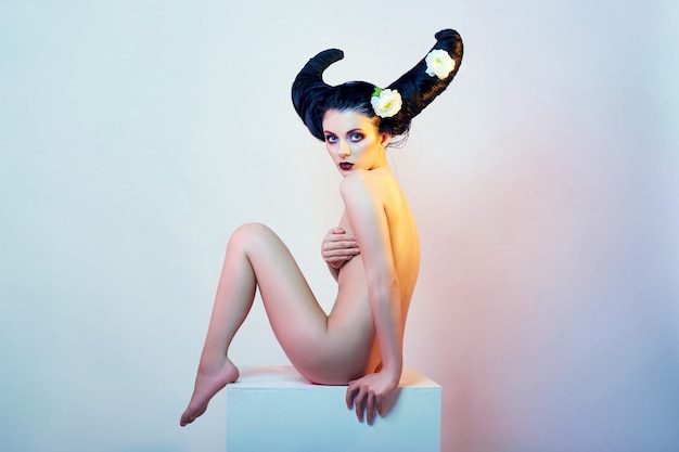 Art fashion Nude girl with hair horns sitting on a white cube. Beautiful brunette devil woman, Horny demon girl. Perfect body and bright makeup