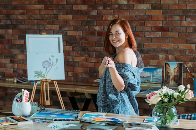 Art hobby. Studio workspace atmosphere. Smiling redhead female posing with paintbrushes and easel