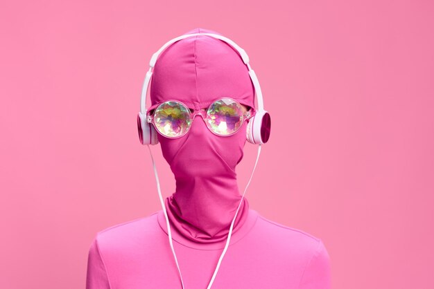 Art portrait of a woman wearing a pink fullface burglar mask with glowing round glasses wearing pink clothes with pink headphones on a pink background looking into the camera high quality photo