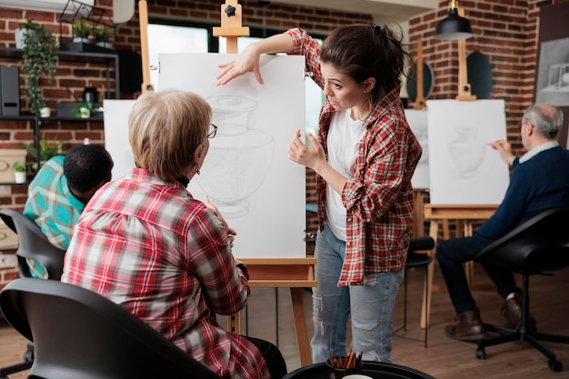 Art teacher helping senior student to develop sketching technique while attending at artistic lesson for personal growth. Multi ethnic group of students drawing vase model on white canvas