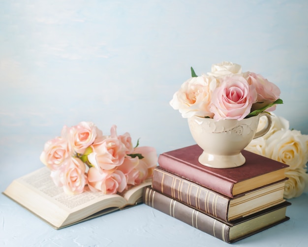 Artificial pink rose flowers in vintage cup with books on blue 