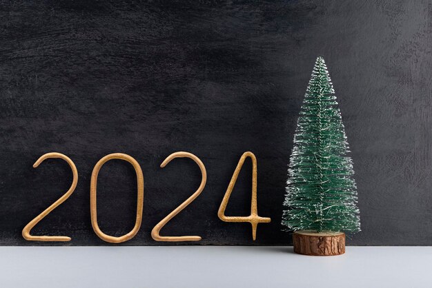 Artificial small Christmas tree on wooden stand and golden number 2024 on black background New year background