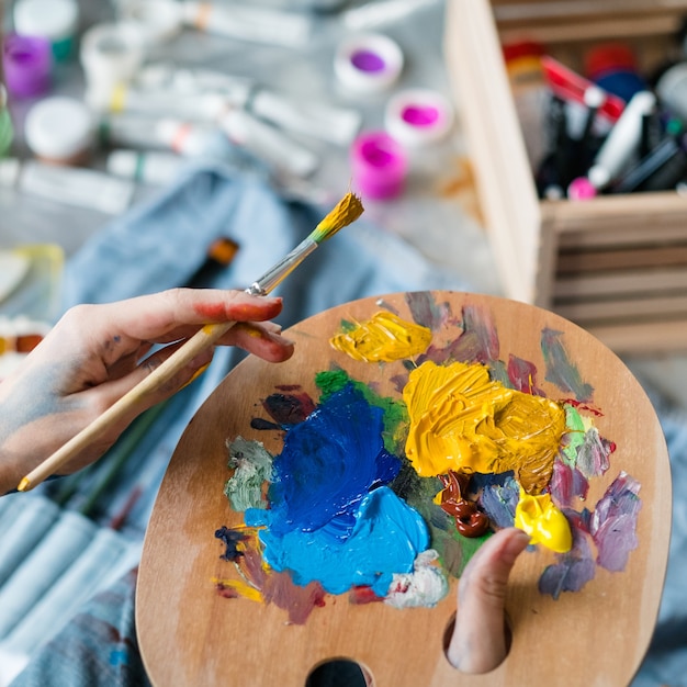 Artist essential tools. closeup of female hands holding wooden palette and paintbrush over blurred paint tubes.