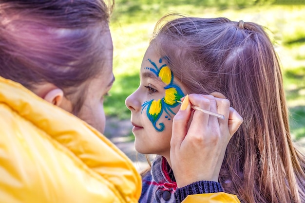 An artist's hand draws a blueyellow flower on the face of a little girl A child with a funny face painting Children's holiday