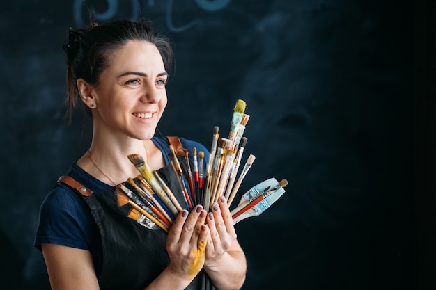 Photo artist supplies. artwork in process. smiling young woman in apron with paintbrushes assortment