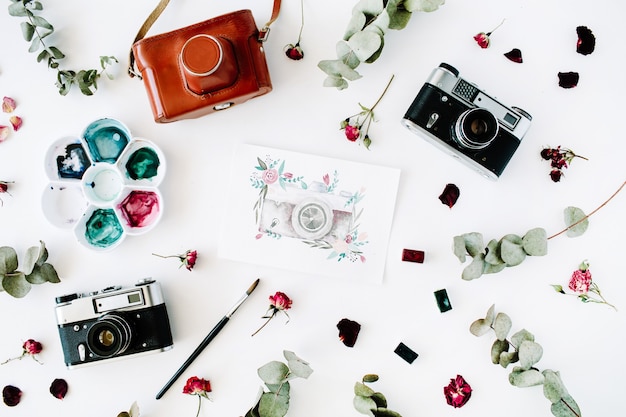 Area di lavoro dell'artista con macchina fotografica retrò vintage e macchina fotografica dipinta ad acquerello, rose rosse ed eucalipto